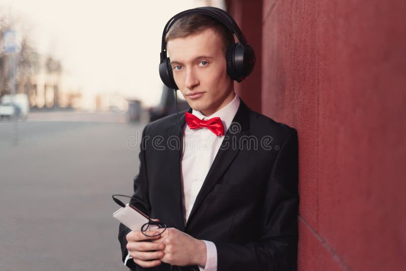Stående av en ung grabb i en svart dräkt och en röd fluga man som lyssnar till musik i stor hörlurar fotografering för bildbyråer Stående av en ung grabb i en svart dräkt och en röd fluga man som lyssnar till musik i stor hörlurar fotografering för bildbyråer