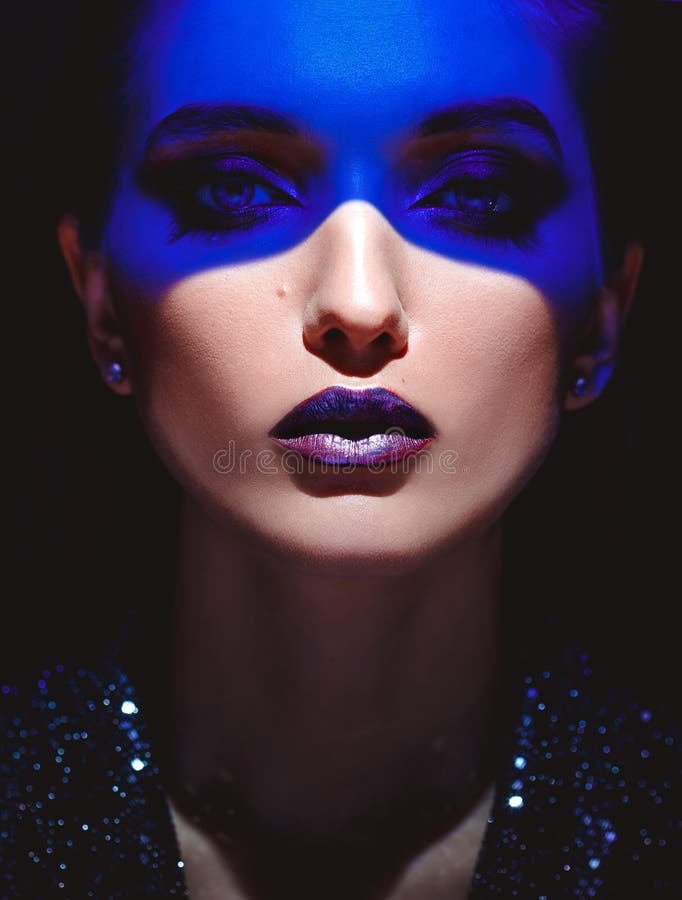 Stående av modeflickan med stilfull makeup och blått neonljus på hennes framsida på den svarta bakgrunden i studion