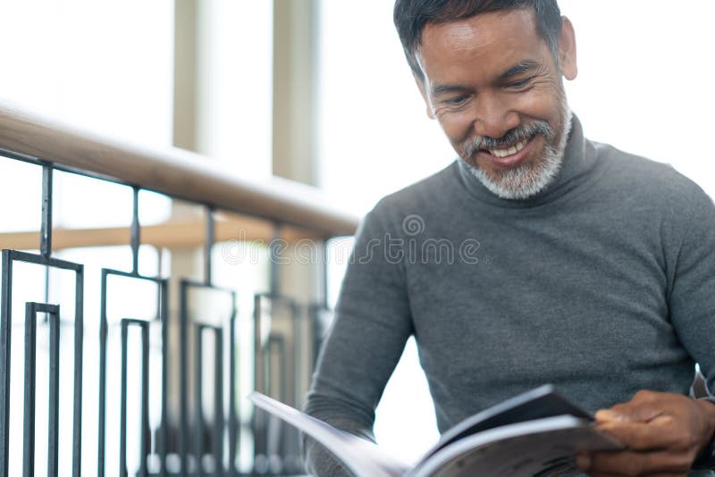 Ståenden av den attraktiva mogna asiatiska mannen avgick med stilfullt kort skäggsammanträde som ler och läser tidskriftböcker