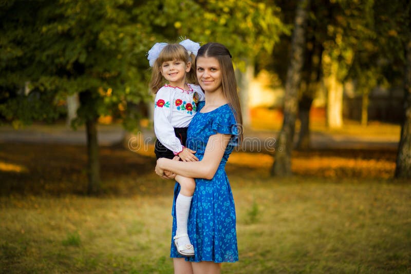 Ståenden av den härliga unga modern som kramar första-väghyveln dottern i festlig skolalikformig på bakgrundshöst, parkerar royaltyfri bild Ståenden av den härliga unga modern som kramar första-väghyveln dottern i festlig skolalikformig på bakgrundshöst, parkerar royaltyfri bild