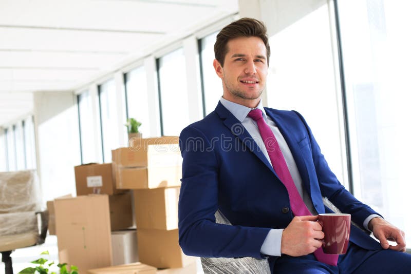 Ståenden av den hållande kaffekoppen för den unga affärsmannen med flyttning boxas i bakgrund på kontoret arkivfoto Ståenden av den hållande kaffekoppen för den unga affärsmannen med flyttning boxas i bakgrund på kontoret arkivfoto