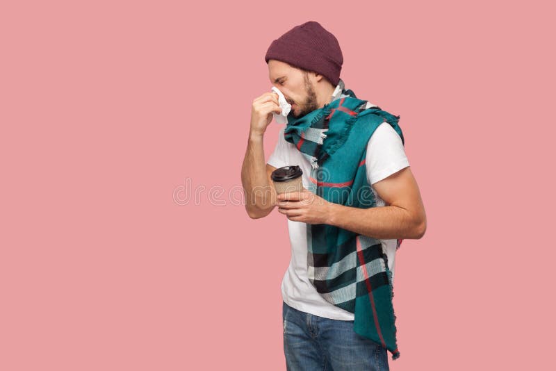 Ståenden av den moderna skäggiga unga hipstermannen för sjukdomen i den vita skjortan och tillfälligt hattanseende, nyser av serv royaltyfria foton Ståenden av den moderna skäggiga unga hipstermannen för sjukdomen i den vita skjortan och tillfälligt hattanseende, nyser av serv royaltyfria foton