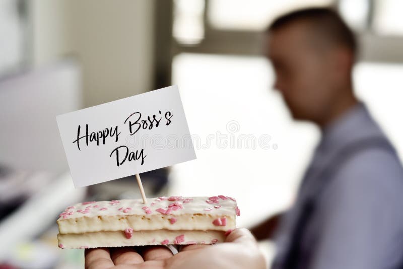 Someone holding oa piece of cake with a signboard with the text happy boss day written in next to a desk, where there is a man working in a computer. Someone holding oa piece of cake with a signboard with the text happy boss day written in next to a desk, where there is a man working in a computer