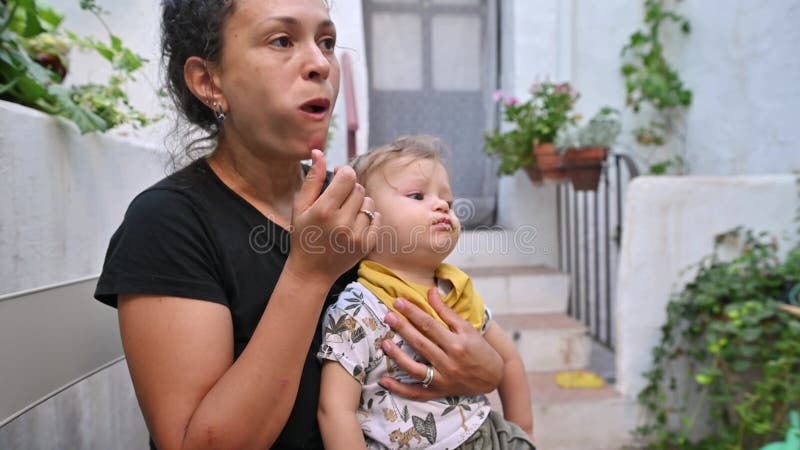 Su madre da de comer a su bebé mientras está sentada al aire libre en un acogedor patio rodeado de exuberante vegetación y un luga almacen de metraje de vídeo
