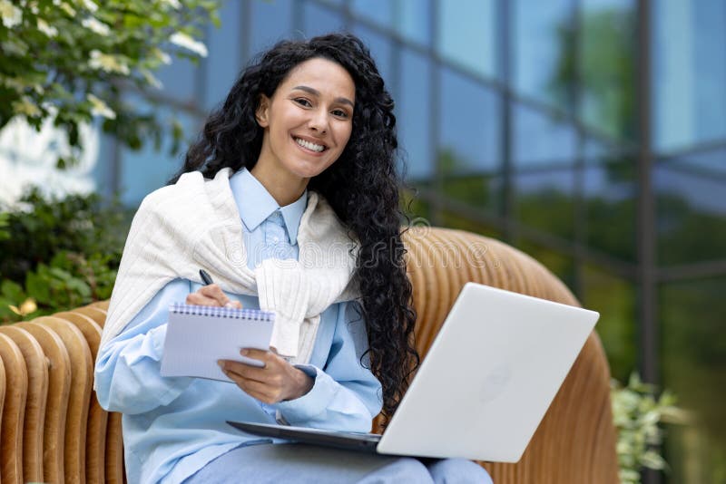 Successful diverse entrepreneur sitting on wooden bench and making notes in daybook while holding computer on laps. Smiling female writing to do list by hand for not skipping important tasks.