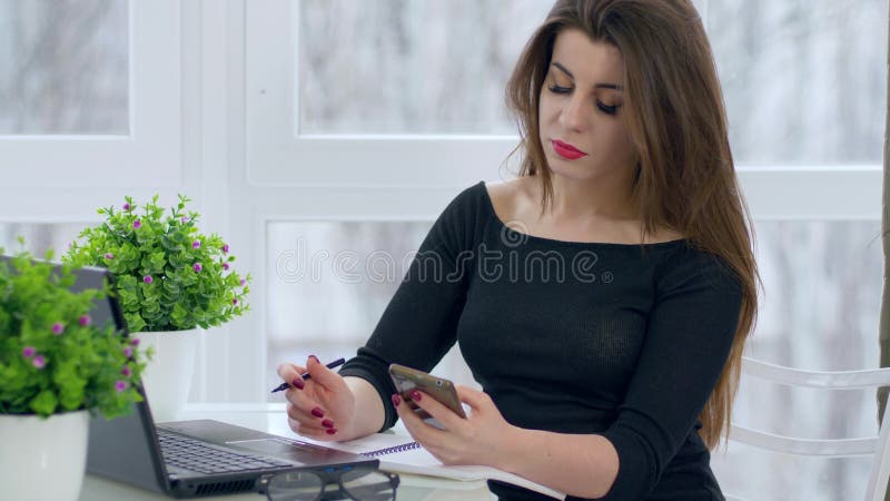 Successful people, long-haired girl uses laptop and mobile at work sitting at desk in light office