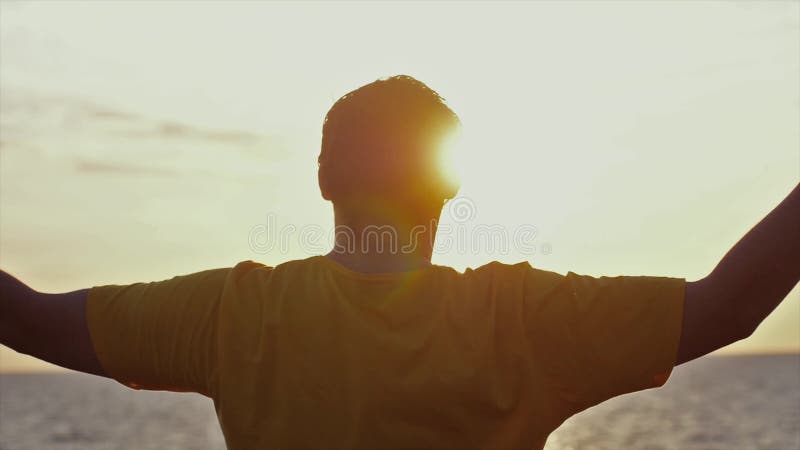 Successful young man is dancing spinning around and raising his hands in slow motion at amazing sunset on the beach