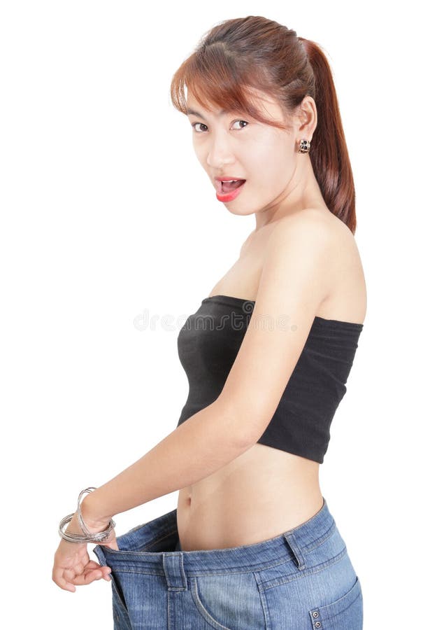Asian slim fit girl or woman gazing amazed at her slimmed down waistline as proven by stretching her loose denim pants after a successful dieting or working out period. Isolated over white. Asian slim fit girl or woman gazing amazed at her slimmed down waistline as proven by stretching her loose denim pants after a successful dieting or working out period. Isolated over white.