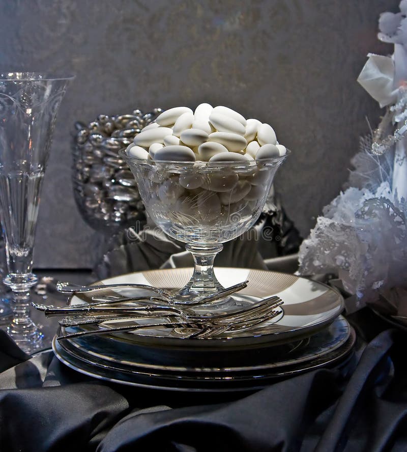 White sugar coated almonds in a crystal glass on top of some plates with forks at a wedding table setting with a dark gray table cloth