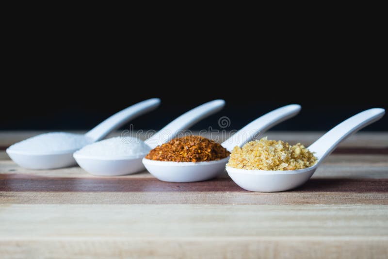 Sugar, salt, pepper and deep fried garlic are placed on a white spoon.And the background is black. This is a garnish.