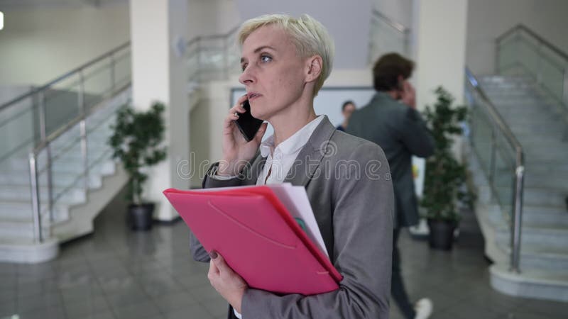 Suivi tir occupé mature caucasienne femme manager à pied parler au téléphone passer des collègues dans le hall de bureau quitter