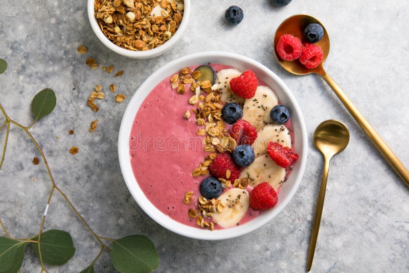 Summer acai smoothie bowls with raspberries, banana, blueberries, and granola on gray concrete background. Breakfast bowl with