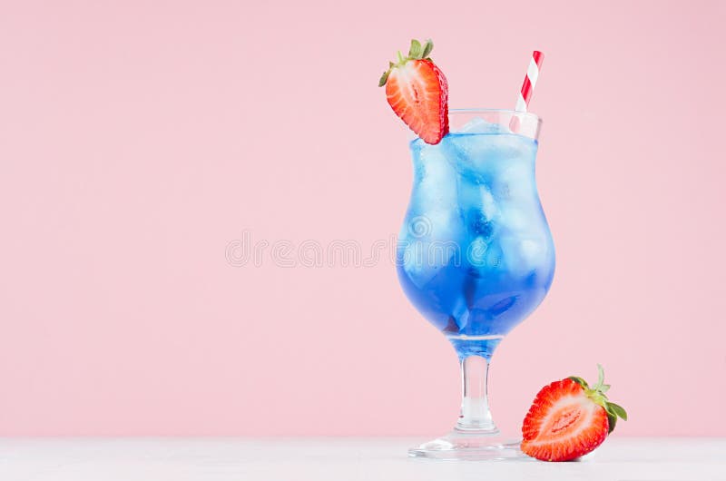 Summer blue alcohol drink with curacao liqueur, ice cubes, strawberry slice, straw in glamour glass on soft light pink background.