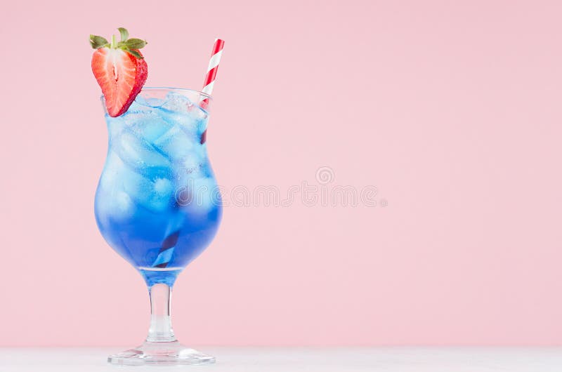 Summer blue alcohol drink with curacao liqueur, ice cubes, strawberry slice, straw in glamour glass on soft light pink background.