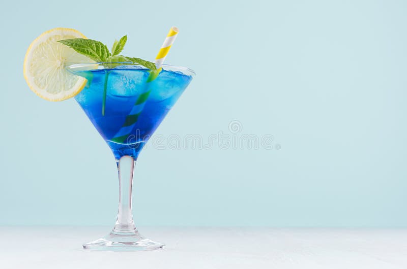 Summer cold blue lagoon drink with ice cubes, lemon slice, yellow straw, green mint in elegant wine glass on pastel mint.