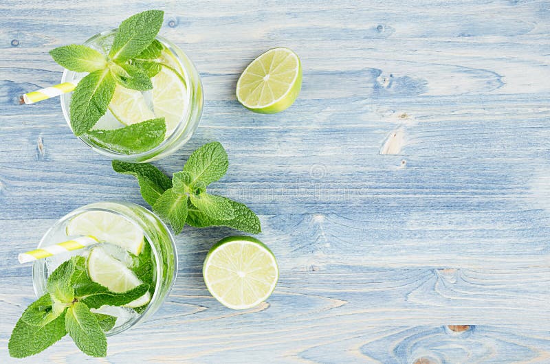 Summer cool lemonade in two wet glasses with mint, lime, ice, straw on soft shabby blue wood board, copy space, top view.