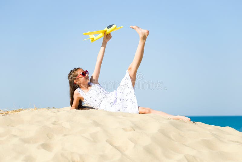 Cute happy little girl wearing sun eyeglasses and a white dress lying on the sandy beach by the sea and playi