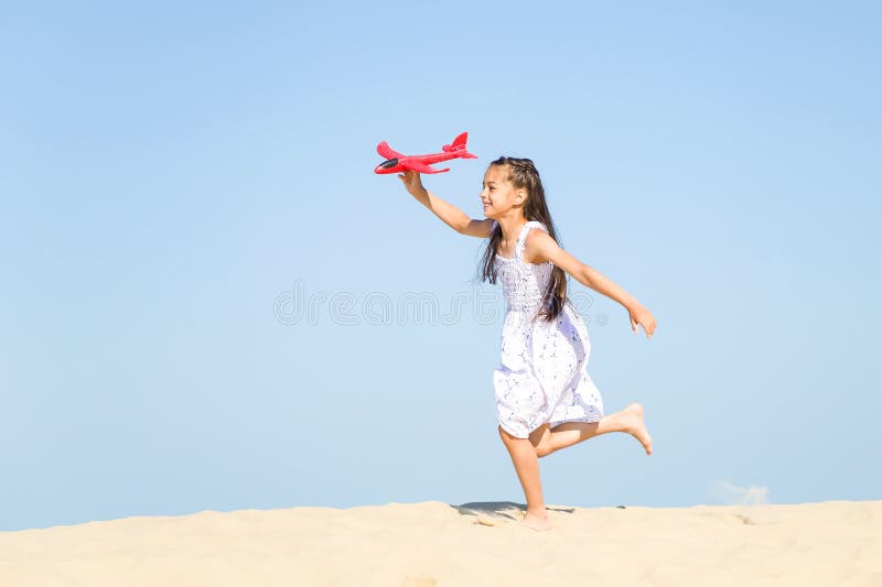 Cute happy little girl wearing a white dress running on the sandy beach by the sea and playing with the red t