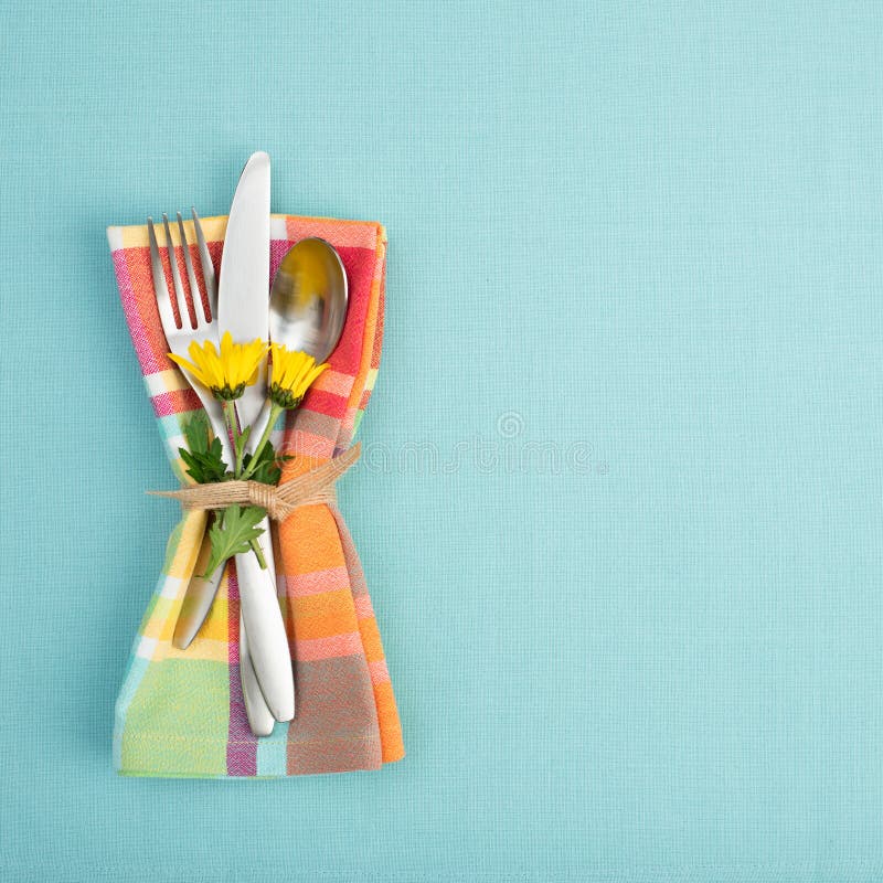 Summer or Spring Table Setting with teal tablecloth, silverware, and multicolor cloth napkin with yellow daisy flowers.  Backgroun