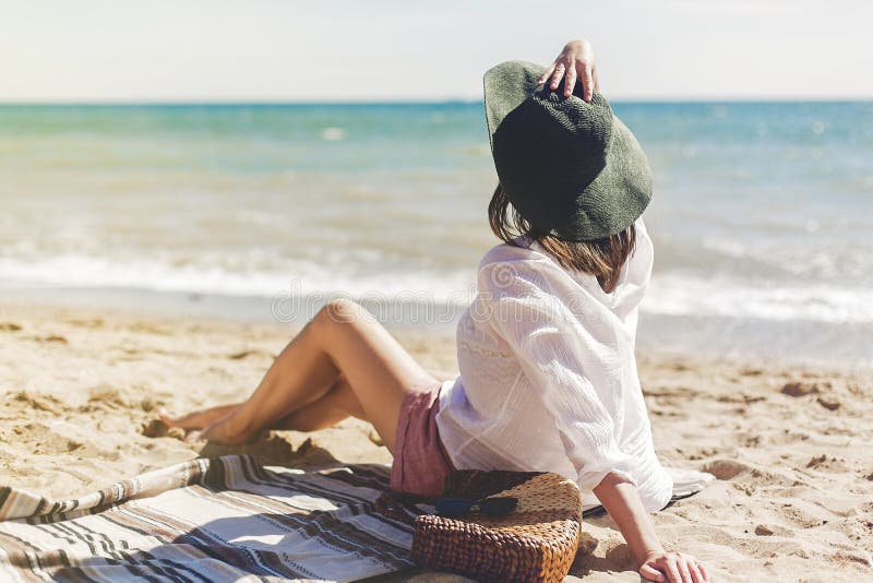 Summer vacation concept. Happy young woman relaxing on beach. Hipster slim girl in white shirt and hat sitting and tanning on