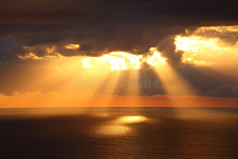 Sunbeams through dark clouds over ocean by golden sunrise