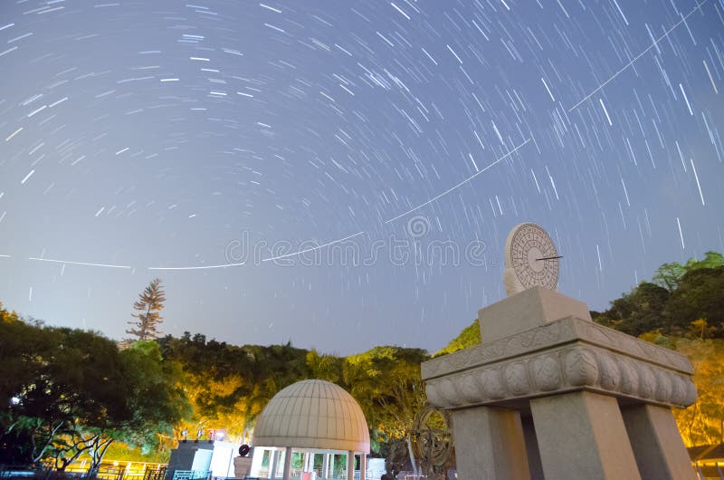 Obrázek rovníkové sluneční hodiny s Čínskými znaky označující čas a star trail a také letadlo stezka v nebi.