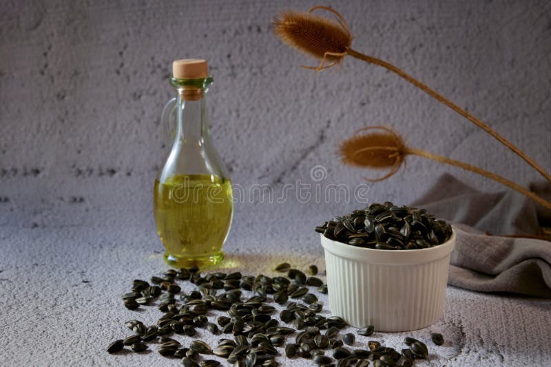 Sunflower seeds in a white ceramic bowl and in bulk on a table with a glass bottle of sunflower oil on a gray background with dry stock image
