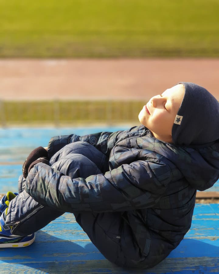 Child sits on a park bench, turns his face to the sun`s rays, enjoys and rejoices in the first warmth royalty free stock photo