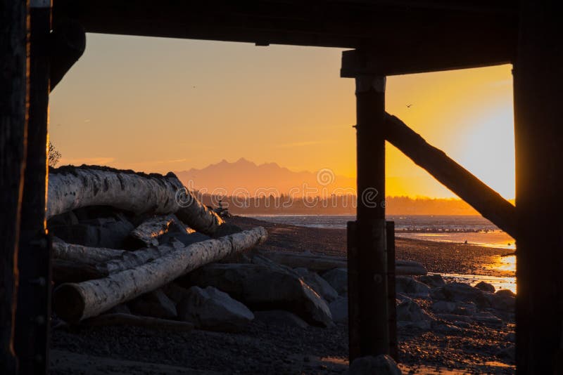 Sunrise is a glorious time on White Rock beach near Vancouver, BC. This shot looks through its wooden pier, past logs piled on beach by winter storms, shallow waters of Semiahmoo Bay, Peace Arch border crossing between Canada and the USA to silhouette of Three Fingers Mountain part of North Cascades range in Washington State. Sunrise is a glorious time on White Rock beach near Vancouver, BC. This shot looks through its wooden pier, past logs piled on beach by winter storms, shallow waters of Semiahmoo Bay, Peace Arch border crossing between Canada and the USA to silhouette of Three Fingers Mountain part of North Cascades range in Washington State.