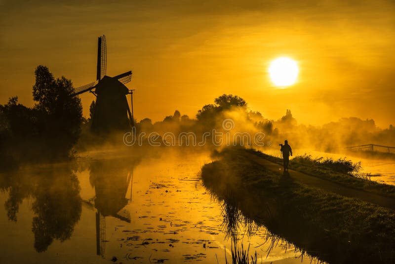 Photographer got out to capture the beautiful early morning and warm sunrise lights heating up the calm water of the canal, around the windmill and the bridge nearby Hazerswoude, Leiden, Netherlands. Photographer got out to capture the beautiful early morning and warm sunrise lights heating up the calm water of the canal, around the windmill and the bridge nearby Hazerswoude, Leiden, Netherlands