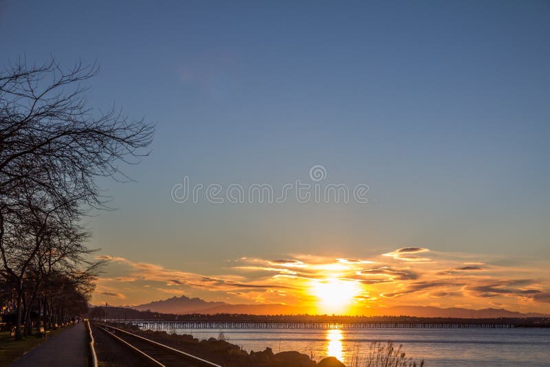 Beautiful sunrise with gold, pink, purple and yellow colors over the North Cascade Mountains of Washington State. Three Fingers Mountain is silhouetted on the horizon and the railway lines along White Rock`s beach shine in the sun. Beautiful sunrise with gold, pink, purple and yellow colors over the North Cascade Mountains of Washington State. Three Fingers Mountain is silhouetted on the horizon and the railway lines along White Rock`s beach shine in the sun.