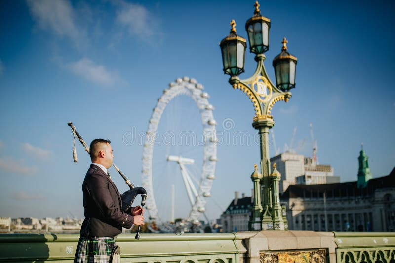 Suonatore di cornamusa che gioca musica scozzese in ponte di Westminster, con London Eye nei precedenti immagini stock Suonatore di cornamusa che gioca musica scozzese in ponte di Westminster, con London Eye nei precedenti immagini stock