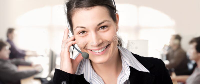 IT-support. Young female customer service representative recieves calls on a headset while colleagues are working on computers in the background IT-support. Young female customer service representative recieves calls on a headset while colleagues are working on computers in the background
