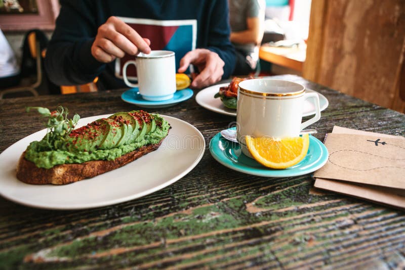 Sur la table est un sandwich utile à veggie ou un pain grillé avec l'avocat et une tasse à côté de thé et d'orange frais Un homme images stock Sur la table est un sandwich utile à veggie ou un pain grillé avec l'avocat et une tasse à côté de thé et d'orange frais Un homme images stock