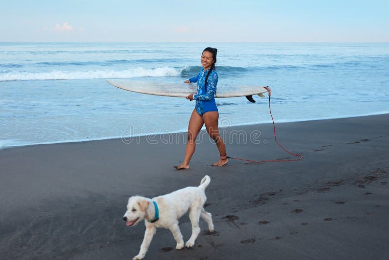 Surfing Girl. Beautiful Woman Walking With Dog On Sandy Beach. Smiling Brunette In Blue Wetsuit Carrying Surfboard. royalty free stock images Surfing Girl. Beautiful Woman Walking With Dog On Sandy Beach. Smiling Brunette In Blue Wetsuit Carrying Surfboard. royalty free stock images