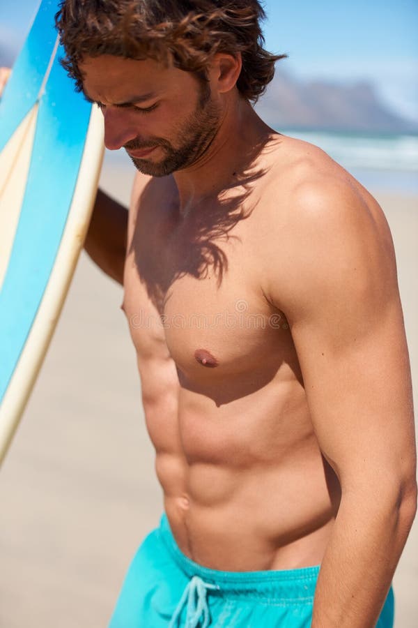 Surfing keeps him in great shape. A young man standing next to his surfboard looking around.