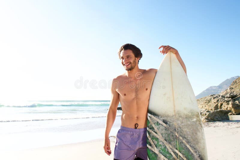 Surfista masculino feliz que está com sua placa na praia fotografia de stock royalty free Surfista masculino feliz que está com sua placa na praia fotografia de stock royalty free