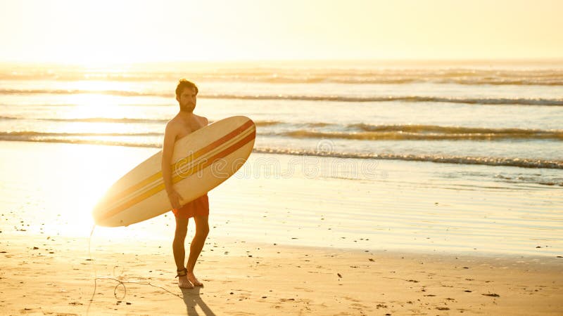 Landscape image of male surfer busy walking on the beach at sunrise while carrying his surfboard under his arm with the ocean waves breaking in the background. Landscape image of male surfer busy walking on the beach at sunrise while carrying his surfboard under his arm with the ocean waves breaking in the background.