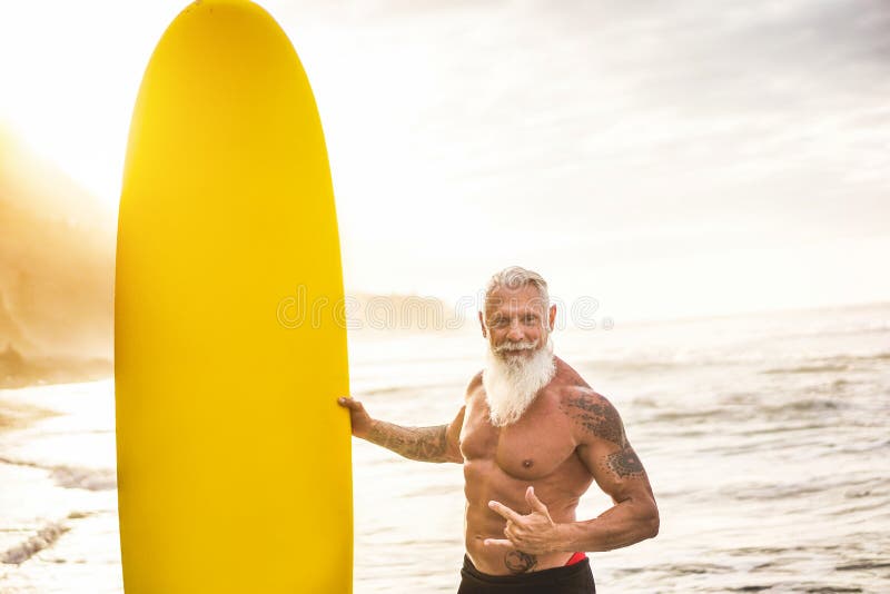 Tattooed senior surfer holding surf board on the beach at sunset - Happy old guy having fun doing extreme sport - Joyful elderly concept. Tattooed senior surfer holding surf board on the beach at sunset - Happy old guy having fun doing extreme sport - Joyful elderly concept