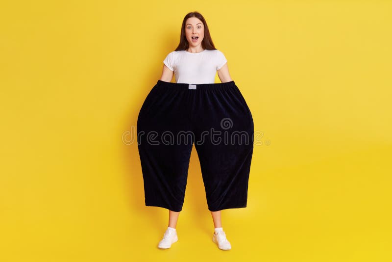 Shocked astonished woman wearing old too big black trousers keeps hands in pants. looks at camera with open mouth and big eyes, has surprised facial expression, posing isolated over yellow background. Shocked astonished woman wearing old too big black trousers keeps hands in pants. looks at camera with open mouth and big eyes, has surprised facial expression, posing isolated over yellow background