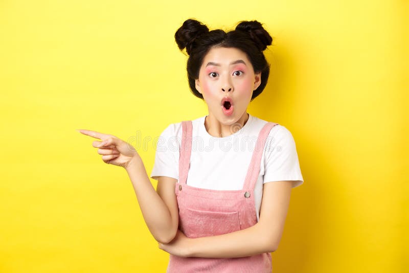 Surprised asian teen girl looking impressed, say wow and pointing finger left, standing with glamour makeup and summer