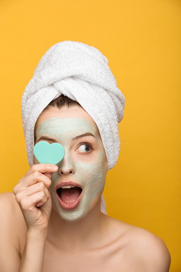 Surprised girl with nourishing facial mask covering eye with heart-shaped cosmetic sponge