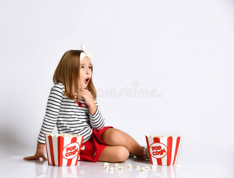 Surprised little girl in a red skirt sits on the floor with a bucket of popcorn on a gray background