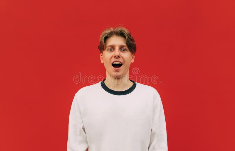 Surprised positive young man in white sweatshirt isolated on red wall background, looking at camera with shocked face