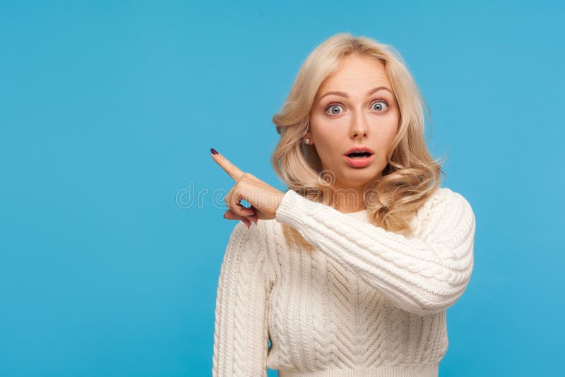 Surprised shocked blond woman in knitted sweater pointing finger away at empty blank for your advertisement, wondered with