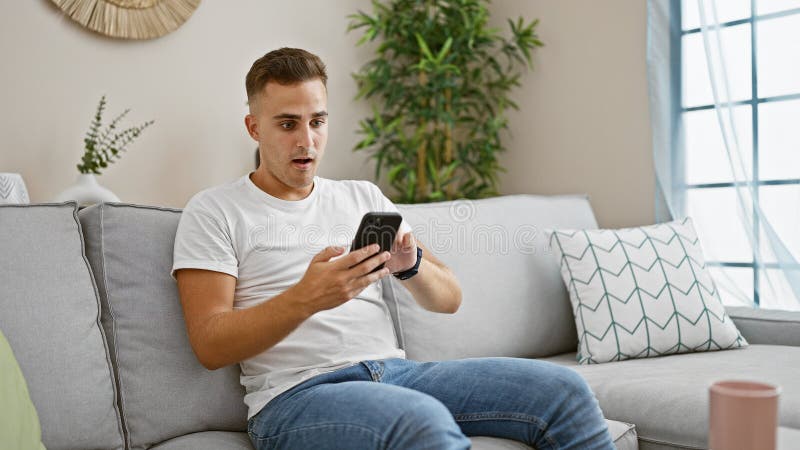Surprised young hispanic man using smartphone in modern living room interior royalty free stock image