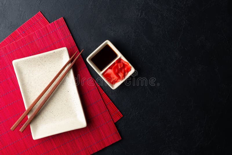 Sushi chopsticks, soy sauce, ginger, red bamboo mat and empty plate on black background.