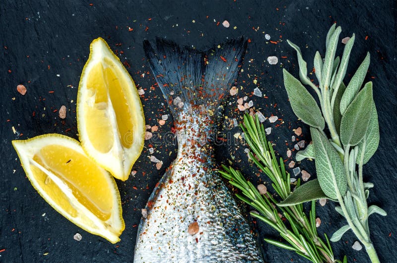 Tail of fresh raw Dorado or sea bream fish on black slate stone board with spices, herbs, lemon and salt. Top view. Tail of fresh raw Dorado or sea bream fish on black slate stone board with spices, herbs, lemon and salt. Top view