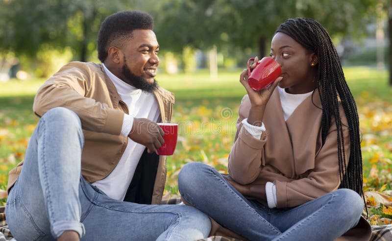 First date outdoors. Black guy and girl sitting on plaid and drinking tea together, dating in autumn park. First date outdoors. Black guy and girl sitting on plaid and drinking tea together, dating in autumn park.