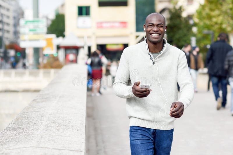 Black young man with a smartphone in his hand in urban background. Young african guy with shaved head wearing casual clothes and white headphones. Black young man with a smartphone in his hand in urban background. Young african guy with shaved head wearing casual clothes and white headphones.